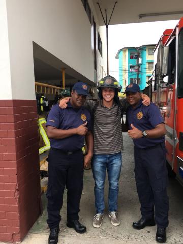 Cameron Wanic taking a photo with two fire fighters and wearing a fire fighter helmet.